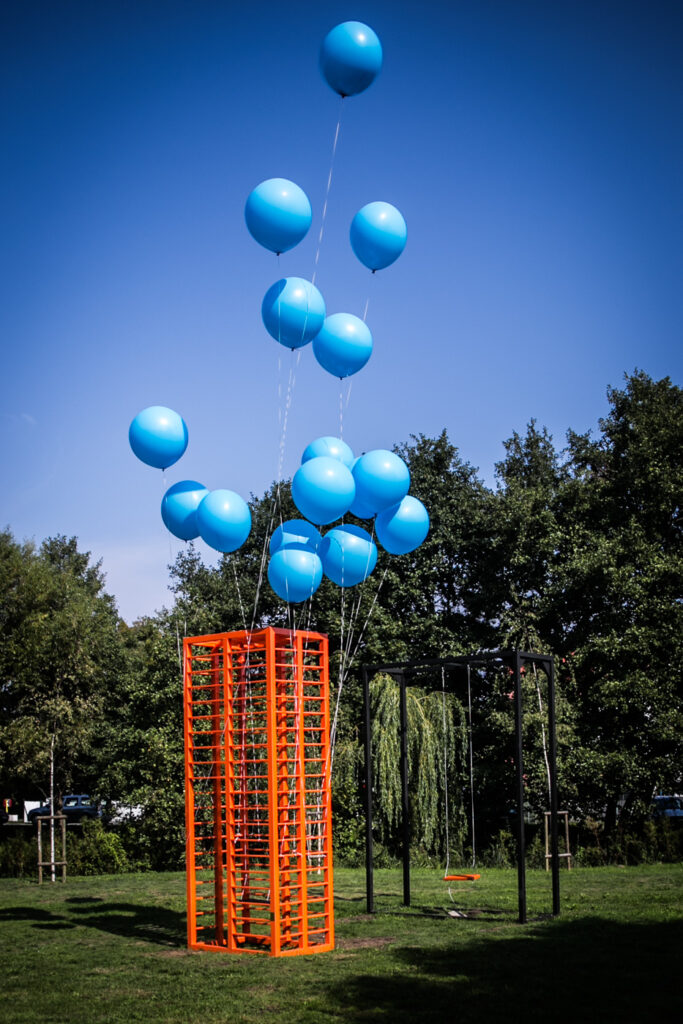 Zdjęcie pionowe przedstawiające abstrakcyjną rzeźbę w przestrzeni parkowej, na której konstrukcję składa się pomarańczowa, metalowy, stojący, wysoki prostopadłościan, którego wnętrze zostało wypełnione wieloma, drobnymi, poziomymi prętami. Do niektórych z nich zostały przywiązane białe liny z unoszącymi się w powietrzu okrągłymi, niebieskimi balonami.