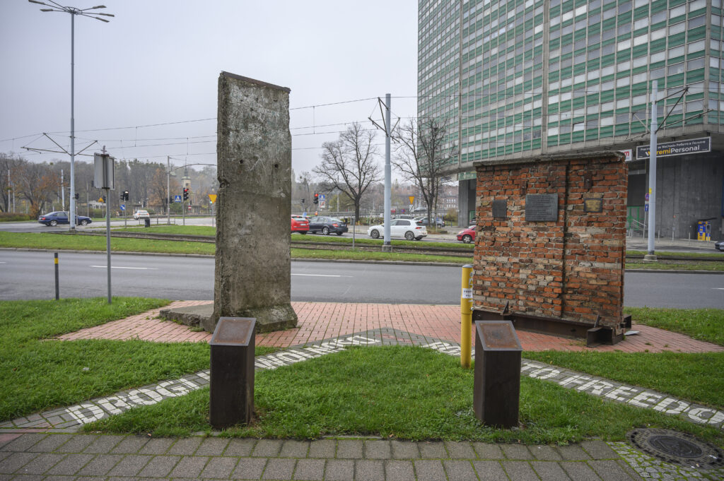 Zdjęcie poziome przedstawiające dwa fragmenty murów: obiekt stojący po prawej stronie wykonany jest z czerwonej cegły, natomiast przeciwległy z betonowej płyty. Ustawione są do siebie wzajemnie pod kątem 45°. Na chodniku u ich podstaw został namalowany biały napis: „Droga do wolności” i „Road to freedom”. W tle widać wielkomiejską zabudowę Gdańska