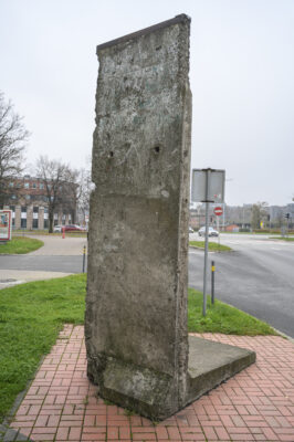 Zdjęcie pionowe przedstawiające fragment ciemnoszarego komponentu wykonanego z żelazobetonu. Ma on formę wysokiego, wąskiego prostokąta. Obiekt znajduje się w przestrzeni o charakterze miejskim.