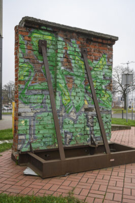 Zdjęcie pionowe przedstawiające fragment muru z czerwonej cegły, który jest wzmocniony dodatkowo metalową konstrukcją przypominającą element stoczniowy. Powierzchnia ściany pokryta jest graffiti w zielonej tonacji o abstrakcyjnie formie. Obiekt znajduje się w przestrzeni o charakterze miejskim.