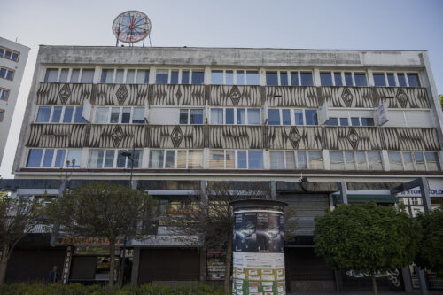 Zdjęcie poziome przedstawiające modernistyczny budynek, którego trzy górne kondygnacje zostały urozmaicone dwoma szerokimi pasami mozaiki. Utrzymane są one w różnych odcieniach szarości i mają charakter geometryczny. Ich kompozycje są zdominowane półkolami oraz rombami, na których zostały umieszczone różne symbole.