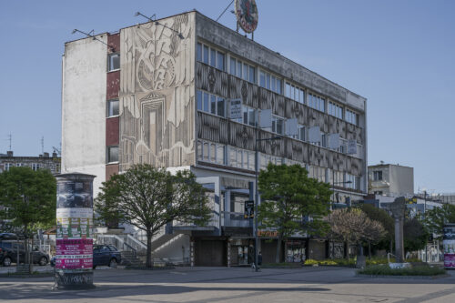 Zdjęcie poziome przedstawiające modernistyczny budynek. Jedna z jego ścian została urozmaicona dwoma szerokimi pasami mozaiki. Utrzymane są one w różnych odcieniach szarości i mają charakter geometryczny. Ich kompozycje są zdominowane półkolami oraz rombami, na których zostały umieszczone różne symbole. Ściana frontalna natomiast została pokryta nowoczesny sgraffito, które jest utrzymane z stylistyce futurystycznej.