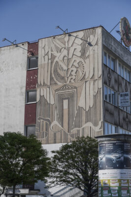 Zdjęcie pionowe przedstawiające modernistyczny budynek, którego ściana frontalna została pokryta nowoczesny sgraffito, które jest utrzymane z stylistyce futurystycznej. Dekoracja utrzymana jest w różnych odcieniach jasnych brązów a jej kompozycja oparta jest na centralnej osi pionowej.