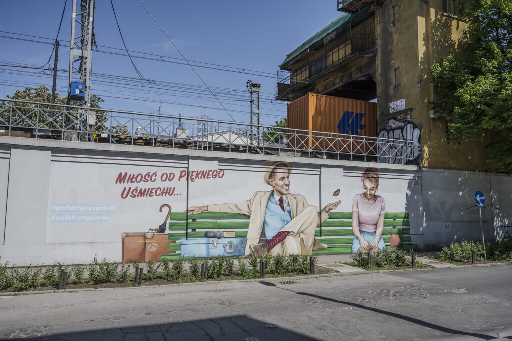 Zdjęcie poziome przedstawiające mural wykonany na części betonowego ogrodzenia w przestrzeni miejskiej, Przedstawia on mężczyznę i kobietę siedzących na drewnianej, zielonej ławce. Ich stroje oraz stylistyka przedstawienia nawiązuje do lat 50. XX wieku. Przedstawione postaci uśmiechają się szeroko, a atmosfera całej ukazanej sceny jest beztroska i radosna. Po lewej stronie znajduje się napis: „Miłość do pięknego uśmiechu…”. Całej przedstawienie utrzymane jest w jasnej i różnobarwnej kolorystyce.