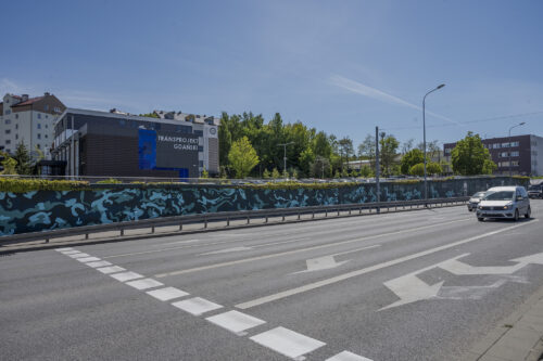 Zdjęcie poziome przedstawiające mural wykonany na części betonowego ogrodzenia ustawionego wzdłuż wielkomiejskiej ulicy. Przedstawienie utrzymane jest w ciemnej, zielono-niebieskiej kolorystyce w różnej tonacji. Jego kompozycja przypomina plamy moro, a niektóre z nich „przeistaczają się” w wilki.