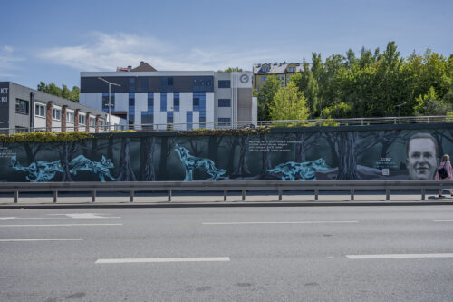 Zdjęcie poziome przedstawiające mural wykonany na części betonowego ogrodzenia ustawionego wzdłuż wielkomiejskiej ulicy. Przedstawienie utrzymane jest w ciemnej, zielono-niebieskiej kolorystyce w różnej tonacji. Na jego kompozycję składają się przedstawienia wilków oraz portretów Żołnierzy Wyklętych wraz z notami informacyjnymi o ich pseudonimach oraz latach życia.