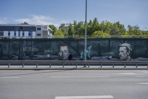 Zdjęcie poziome przedstawiające mural wykonany na części betonowego ogrodzenia ustawionego wzdłuż wielkomiejskiej ulicy. Przedstawienie utrzymane jest w ciemnej, zielono-niebieskiej kolorystyce w różnej tonacji. Na jego kompozycję składają się przedstawienia wilków oraz portretów Żołnierzy Wyklętych wraz z notami informacyjnymi o ich pseudonimach oraz latach życia.