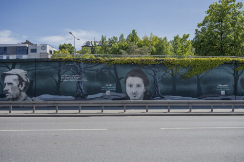 Zdjęcie poziome przedstawiające mural wykonany na części betonowego ogrodzenia ustawionego wzdłuż wielkomiejskiej ulicy. Przedstawienie utrzymane jest w ciemnej, zielono-niebieskiej kolorystyce w różnej tonacji. Na jego kompozycję składają się przedstawienia wilków oraz portretów Żołnierzy Wyklętych wraz z notami informacyjnymi o ich pseudonimach oraz latach życia.