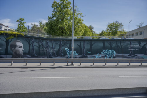 Zdjęcie poziome przedstawiające mural wykonany na części betonowego ogrodzenia ustawionego wzdłuż wielkomiejskiej ulicy. Przedstawienie utrzymane jest w ciemnej, zielono-niebieskiej kolorystyce w różnej tonacji. Na jego kompozycję składają się przedstawienia wilków oraz portretów Żołnierzy Wyklętych wraz z notami informacyjnymi o ich pseudonimach oraz latach życia.