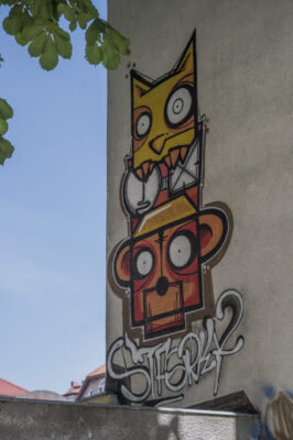 Zdjęcie pionowe ze zbliżeniem na mural wykonany na jednej ze ścian zabytkowej kamienicy. Przedstawiono na nim dwa ssaki, prawdopodobnie małpę i kota, w komiksowej, zantropomorfizowanej konwencji. Zwierzęta ustawione są jeden na drugim, co przypomina układ totemiczny. Mural utrzymany jest w intensywnej żółto-pomarańczowej kolorystyce z białymi elementami oraz podpisem artysty.