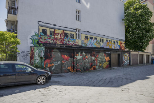 Zdjęcie poziome ukazujące przestrzeń miejskiego blokowiska. Charakterystyczne garaże dla osiedli mieszkaniowych zostały pokryte wieloma muralami o różnej stylistyce i charakterze. Można odnaleźć w tym przypadku przedstawienia portretowe – prawie, że realistyczne oraz bardziej komiksowe. Kolorystyka jest bardzo zróżnicowana: od intensywnych żółcieni, pomarańczy i błękitów do różnych tonacji brązów i czerni.