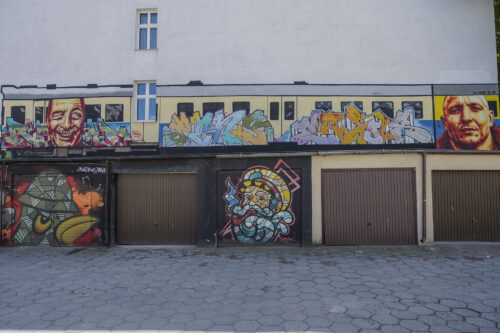 Zdjęcie poziome ukazujące przestrzeń miejskiego blokowiska. Charakterystyczne garaże dla osiedli mieszkaniowych zostały pokryte wieloma muralami o różnej stylistyce i charakterze. Można odnaleźć w tym przypadku przedstawienia portretowe – prawie, że realistyczne oraz bardziej komiksowe. Kolorystyka jest bardzo zróżnicowana: od intensywnych żółcieni, pomarańczy i błękitów do różnych tonacji brązów i czerni.