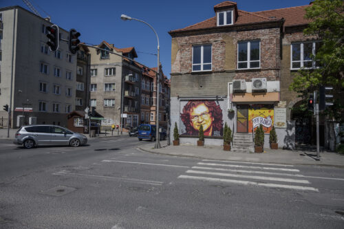 Zdjęcie poziome przedstawiające mural wykonany na jednej ze ścian zabytkowej kamienicy miejskiej. Przedstawienie ma charakter portretowy, na którym znajduje się mężczyzna w średnim wieku. Ma on włosy sięgające ramion a także nosi okulary w wąskiej oprawie. Mural utrzymany jest w ciepłej pomarańczowo-żółto-czerwonej kolorystyce. W prawym, dolnym rogu znajdują się białe litery układające się w napis: „Dzięki Panie Zbyszku”.