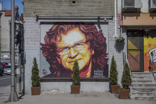 Zdjęcie poziome przedstawiające mural wykonany na jednej ze ścian zabytkowej kamienicy miejskiej. Przedstawienie ma charakter portretowy, na którym znajduje się mężczyzna w średnim wieku. Ma on włosy sięgające ramion a także nosi okulary w wąskiej oprawie. Mural utrzymany jest w ciepłej pomarańczowo-żółto-czerwonej kolorystyce. W prawym, dolnym rogu znajdują się białe litery układające się w napis: „Dzięki Panie Zbyszku”.