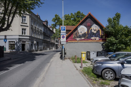 Zdjęcie poziome przedstawiające mural wykonany na jednej ze ścian zabytkowego budynku w przestrzeni miejskiej. Zostały na nim przedstawione dwie kukiełkowe postaci ludzkie z „Muppet Show”. Mężczyźni w podeszłym wieku ubrani są w garnitury oraz białe koszule, i zostali przedstawieni na eleganckich krzesłach z czerwonym obiciem. Postać znajdująca się po prawej stronie ma zgryźliwy wyraz twarzy, natomiast jego towarzysz sprawia wrażenie spokojnego i cichego.