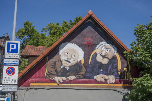 Zdjęcie poziome przedstawiające mural wykonany na jednej ze ścian zabytkowego budynku w przestrzeni miejskiej. Zostały na nim przedstawione dwie kukiełkowe postaci ludzkie z „Muppet Show”. Mężczyźni w podeszłym wieku ubrani są w garnitury oraz białe koszule, i zostali przedstawieni na eleganckich krzesłach z czerwonym obiciem. Postać znajdująca się po prawej stronie ma zgryźliwy wyraz twarzy, natomiast jego towarzysz sprawia wrażenie spokojnego i cichego.