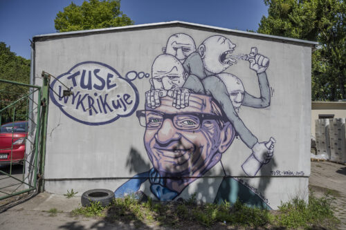 Zdjęcie poziome przedstawiające mural wykonany na jednej ze ścian garażu miejskiego. Przedstawia on mężczyznę w średnim wieku z cwanym wyrazem twarzy, a jego przedstawienie zostało wykonane w bardzo realistyczny sposób. Kontrastuje to silnie z czterema komiksowymi postaci, które wydostają się z jego głowy. Przy jednej z nich znajduje się chmurka myślowa z napisem: „Tuse wykrikuje”.