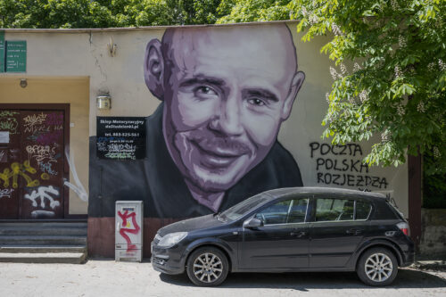 Zdjęcie poziome przedstawiające mural wykonany na ścianie budynku znajdującego się w przestrzeni miejskiej. Przedstawia on młodego mężczyznę ukazanego w sposób realistyczny. Ma on ogoloną na łyso głowę oraz nosi niewielki zarost oraz ubrany jest w czarną koszulę. Mężczyzna uśmiecha się szczerze do widza i nawiązuje z nim kontakt wzrokowy.