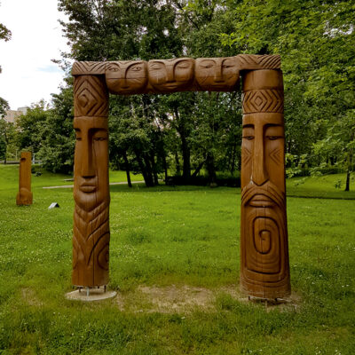 Zdjęcie poziome przedstawiające rzeźbę abstrakcyjną w przestrzeni parkowej. Rzeźba została wykonana w pamarańczowo-brązowym drewnie. Jej konstrukcja przypomina kwadratową ramę zbudowaną z trzech osobnych pni. Ich powierzchnie są pokryte płaskorzeźbami, które swoją stylistyką nawiązują do sztuki Azji Południowo-Wschodniej czy Oceanii.