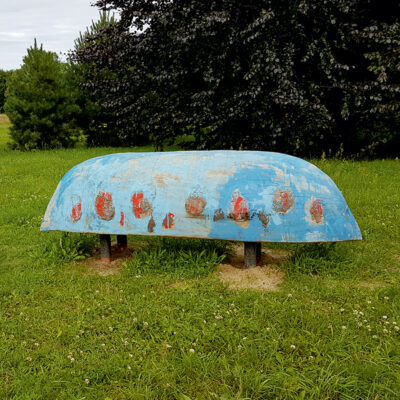 Zdjęcie przedstawiające instalację artystyczną w przestrzeni parkowej. Przypomina ona odwróconą do góry dnem niebieską łódź, która spoczywa na dwóch, czarnych stelażach. Jej powierzchnia jest stosunkowo zniszczona i zaczyna pojawiać się na niej wyraźny proces korozji.