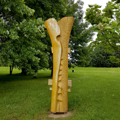 Zdjęcie przedstawiające rzeźbę abstrakcyjną w przestrzeni parkowej. Rzeźba została wykonana w jasnobrązowym drewnie i przedstawia dwie przenikające się, kontrastujące masy. Jednak z nich ma gładką powierzchnie, w przeciwieństwie do drugiej, która ma powierzchnie chrapowatą.