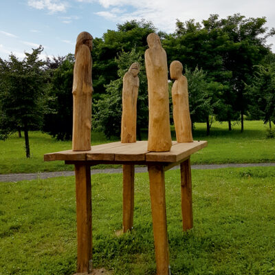 Zdjęcie przedstawiające instalację artystyczną w przestrzeni parkowej. Na jej konstrukcje składa się wysoki, drewniany podest, który swoim wyglądem przypomina wiejski stołek. Na jego rogach zostały ustawione cztery figury ludzkie, które zostały opracowane w bardzo schematyczny a wręcz minimalistycznym sposób.