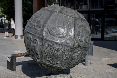Zdjęcie poziome przedstawiające rzeźbę w przestrzeni miejskiej. Przypomina ona globus, na którego powierzchni wyryte zostały przedstawienia zwierzęce a także sceny rodzajowe. Rzeźba podobnie jak globus jest delikatnie pochylona wzdłuż swojej osi środkowej.