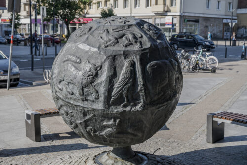 Zdjęcie poziome przedstawiające rzeźbę w przestrzeni miejskiej. Przypomina ona globus, na którego powierzchni wyryte zostały przedstawienia zwierzęce a także sceny rodzajowe. Rzeźba podobnie jak globus jest delikatnie pochylona wzdłuż swojej osi środkowej.