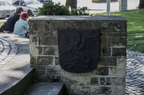 Zdjęcie poziome przedstawiające zbliżenie na wmontowaną w ścianę niewielkiego muru tablicę przypominającą swoim kształtem tarczę herbową hiszpańską. Na jej polu został przedstawiony stający na tylnych łapach gryf w koronie.