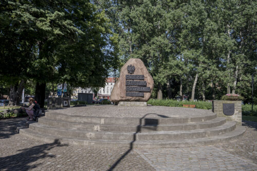 Zdjęcie poziome przedstawiające jasnoróżową, jednolitą, monolityczną skałę, która pełni funkcję upamiętniające. Została ona ustawiona na niskim, prawie płaskim postumencie, a na jednej z jej ścian został umieszczony napis: „Polskie Państwo Podziemne 1939-1945”. Pomnik znajduje się na lekkim podwyższeniu, do którego prowadzą czterostopniowe schody.