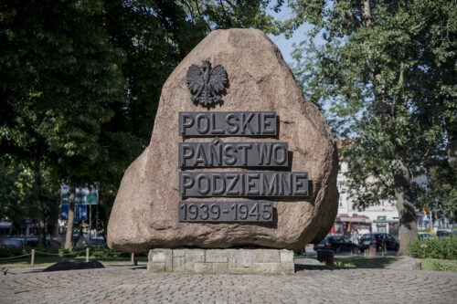 Zdjęcie poziome przedstawiające jasnoróżową, jednolitą, monolityczną skałę, która pełni funkcję upamiętniające. Została ona ustawiona na niskim, prawie płaskim postumencie, a na jednej z jej ścian został umieszczony napis: „Polskie Państwo Podziemne 1939-1945”.