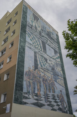 Zdjęcie pionowe wykonane z ukosa przedstawiające ścianę boczną wielomieszkaniowego budynku, która została praktycznie w całości pokryta muralem. Jego kompozycja przedstawia abstrakcyjną i odrealnioną scenę przedstawiającą m.in. żołnierzy, tygrysa i szachownice. Całość utrzymana jest w zielono-niebieskiej tonacji barwnej i przypomina marę senną lub malarstwo Salvadora Dali’ego