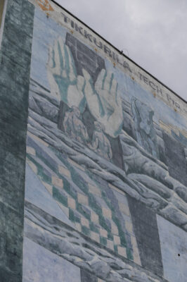 Zdjęcie pionowe wykonane z ukosa przedstawiające fragment muralu pokrywający ścianę boczną wielomieszkaniowego budynku. Jego kompozycja przedstawia abstrakcyjną i odrealnioną scenę przedstawiającą m.in. ludzi, przeskalowane dłonie i geometryczne konstrukcje. Całość utrzymana jest w zielono-niebieskiej tonacji barwnej i przypomina marę senną lub malarstwo Salvadora Dali’ego