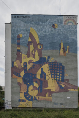Zdjęcie pionowe przedstawiające mural pokrywający w całości ścianę boczną wielomieszkaniowego budynku. Kompozycja muralu oparta jest na wielu figurach geometrycznych, które układają się w abstrakcyjną formę przypominającą strukturę miasta. Wszystkie elementy utrzymane są w żółto-niebiesko-ciemnoczerwonej kolorystyce i umieszczone zostały na szaro-granatowym tle o stłumionej tonacji.
