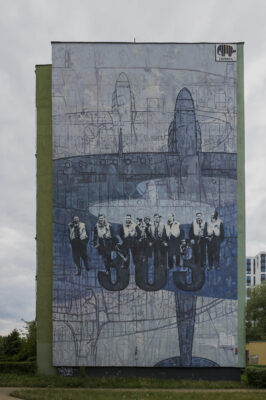 Zdjęcie pionowe przedstawiające mural poświęcony pamięci Dywizjonu 303, który pokrywa całą ścianę boczną wielomieszkaniowego budynku. Kompozycja utrzymana jest w błękitno-granatowej kolorystyce zaś postaci lotników zostały oddane w sposób realistyczny.