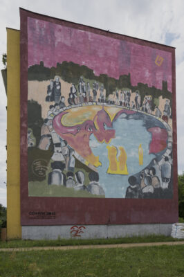 Zdjęcie pionowe przedstawiające mural pokrywający całą ścianę boczną wielomieszkaniowego budynku. Na kompozycje muralu składają się przedstawienia ludzkie, jak także elementy abstrakcyjne, zaś wszystko utrzymane jest w intensywnej, wielobarwnej kolorystyce.