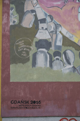 Zdjęcie pionowe z zbliżeniem na fragment muralu pokrywający w całości ścianę boczną wielomieszkaniowego budynku. Na zbliżeniu znajduje się odwrócona plecami do widza ludzka postać, nad której głowa znajduje się chmurka dialogowa, wewnątrz której znajduje się informacja o autorze i realizacji.