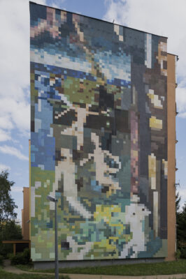 Zdjęcie pionowe przedstawiające mural pokrywający całą ścianę boczną wielomieszkaniowego budynku. Całe przedstawienie ukazane na muralu jest silnie rozpikselowane, dlatego też trudno określić co ono przedstawia. Jego kompozycja utrzymana jest w różnych odcieniach niebieskiego, zielonego, brązu oraz żółcieni.