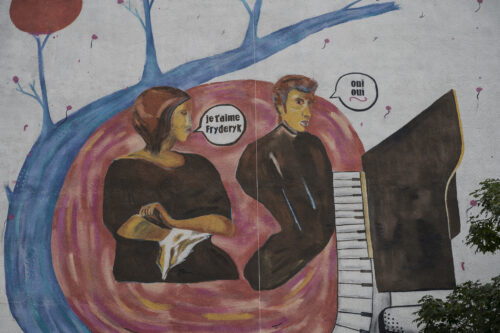 Zdjęcie poziome przedstawiające fragment muralu pokrywającego ścianę boczną budynku wielomieszkaniowego. Przedstawia on Fryderyka Chopina oraz George Sand, którzy zostali ukazani w konwencji komiksowej. Cała kompozycja utrzymana jest w niebiesko-brązowo-czerwonej kolorystyce.