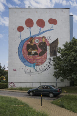 Zdjęcie pionowe przedstawiające mural pokrywający ścianę boczną budynku wielomieszkaniowego. Przedstawia on Fryderyka Chopina oraz George Sand, którzy zostali ukazani w konwencji komiksowej, delikatnie odrealnionej i zniekształconej. Cała kompozycja utrzymana jest w niebiesko-brązowo-czerwonej kolorystyce z biało-czarnymi elementami.