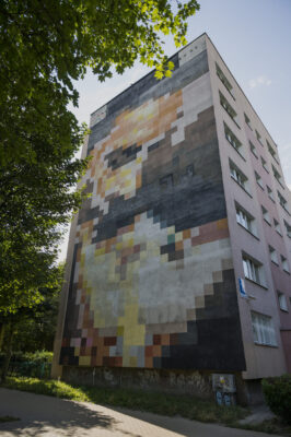 Zdjęcie pionowe przedstawiające mural pokrywający całą ścianę boczną wielomieszkaniowego budynku. Kompozycja muralu przypomina kadr słynnej sceny pocałunku Rhetta Butlera i Scarlett O'Hary z filmu Przeminęło z wiatrem. a całość utrzymana jest w ciepłej, żółto-pomarańczowo-brązowej kolorystyce.