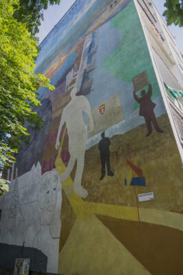Zdjęcie pionowe przedstawiające fragment muralu pokrywającego całą ścianę boczną wielomieszkaniowego budynku. Na jego kompozycje składa się kilka przedstawień ludzkich ukazanych w uproszczony sposób. Zostały one umieszczone na schematycznie przedstawionym krajobrazie. Całość kompozycji utrzymana jest w wielobarwnej, przygaszonej kolorystyce.
