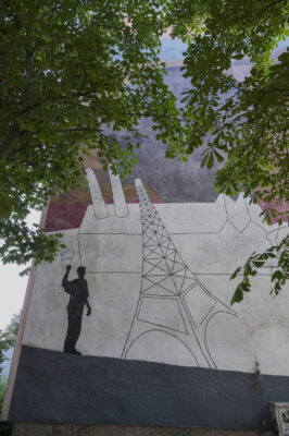 Zdjęcie pionowe przedstawiające fragment muralu pokrywającego całą ścianę boczną wielomieszkaniowego budynku. Na jego kompozycje składa się przedstawienie ludzkie ukazane w uproszczony sposób. Zostało ono umieszczone na schematycznie przedstawionym krajobrazie z charakterystycznym kształtem przypominającym Wieżę Eiffla. Całość kompozycji utrzymana jest w wielobarwnej, przygaszonej kolorystyce.
