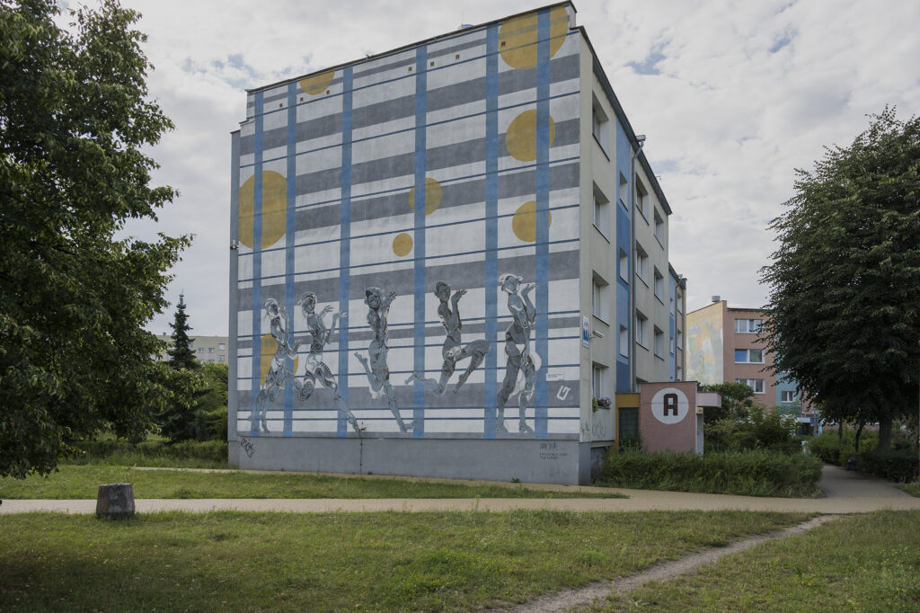 Zdjęcie poziome przedstawiające mural pokrywający całą ścianę boczną wielomieszkaniowego budynku. Przedstawia on pięć kobiet, które zostały ukazane w sposób odrealniony i zniekształcony. Znajdują się one na tle niebiesko-szaro-białej kratki, która jest dodatkowo urozmaicona żółtymi okręgami.
