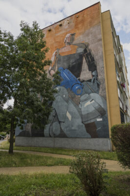 Zdjęcie pionowe przedstawiające mural pokrywający ścianę boczną budynku wielomieszkaniowego. Na pomarańczowym tle umieszczono atletycznie zbudowanego robota. Całość utrzymana jest w pomarańczowo-szarej kolorystyce, z silną niebieską dominantą, zaś przedstawiona postać nasuwa silne skojarzenia ze sztuką komisową.