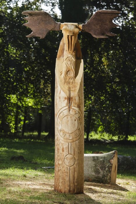 Zdjęcie pionowe przedstawiające drewnianą rzeźbę, przypominającą swoim wyglądem indiański totem. Na jej powierzchni zostały wyryte pogańskie symbole zaś na szczecie umieszczono element nasuwający skojarzenia z czaszką łosia z porożami o uproszczonej formie.