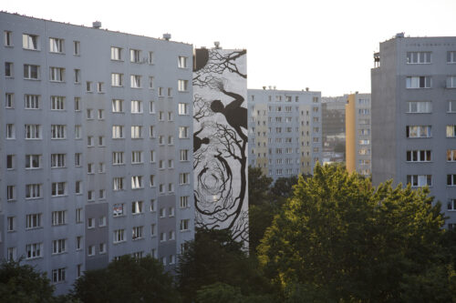 Zdjęcie poziome przedstawiające boczną ścianę jednego z wielokondygnacyjnych budynków mieszkalnych, której powierzchnia została pokryta monochromatycznym muralem. Przedstawia on syntetycznie ukazane figury ludzkie, z których kończyn oraz głów wyrastają pnącza roślinne. Układają się one w formie wiru i pokrywają równomiernie całą powierzchnie ściany.