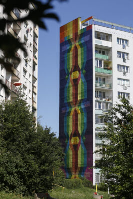 Zdjęcie pionowe przedstawiające boczną ścianę wielokondygnacyjnego budynku mieszkalnego, której powierzchnia została pokryta muralem o abstrakcyjnym charakterze. Jego kompozycja jest symetryczna, osadzona wzdłuż osi pionowej, a swoim wyglądem przypomina zapleciony, długi warkocz. Krawędzie figur, które budują kompozycję muralu są rozmyte, przez co pojawia się wrażenie wzajemnego ich przenikania. Kolorystka malowidła jest intensywna, utrzymana w raczej zimnej niebiesko-fioletowej tonacji, jednak występują również żółte, czerwone i pomarańczowe kontrapunkty kolorystyczne.