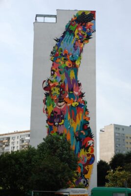 Zdjęcie pionowe przedstawiające boczną ścianę wielokondygnacyjnego budynku mieszkalnego, której powierzchnia została pokryta wielkobarwnym muralem. Jego kompozycja jest wielkoelementowa, której postaci są odrealnione i przedstawione raczej syntetycznie. Mural utrzymany jest wielobarwnej, czystej i nasyconej kolorystyce. Przypomina on kolorową falę wychodzącą z prawego, górnego rogu ściany, która rozlewa się równomiernie ku dołowi.