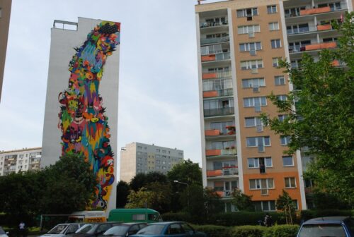 Zdjęcie poziome przedstawiające boczną ścianę jednego z wielokondygnacyjnych budynków mieszkalnych. Jej powierzchnia została pokryta wielkobarwnym muralem. Jego kompozycja jest wielkoelementowa, której postaci są odrealnione i przedstawione raczej syntetycznie. Mural utrzymany jest wielobarwnej, czystej i nasyconej kolorystyce. Przypomina on kolorową falę wychodzącą z prawego, górnego rogu ściany, która rozlewa się równomiernie ku dołowi.